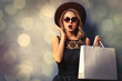 © Masson - Portrait of a young style girl in black dress and hat with shopping bags on gray background with bokeh