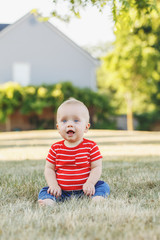 Wall Mural - Cute adorable little Caucasian baby boy in red t-shirt and pants sitting in field meadow outside. Little happy smiling child in summer park on sunset. Lovely charming male infant having fun.