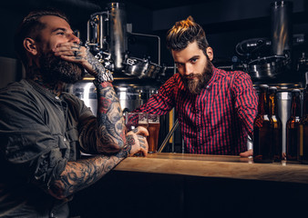 Tattooed male with stylish beard and hair holds pint of craft beer sitting at the bar counter in the indie brewery.