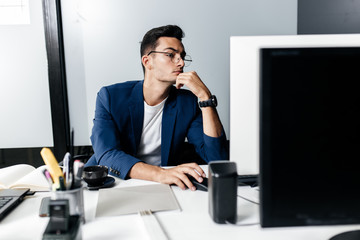 Young man architect in glasses dressed in a business suit sits at a desk in front of a computer in the office