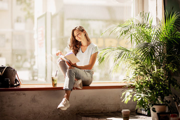 A pretty slim young girl with long hair,wearing casual outfit,sit on the windowsill  and reads a book in a cozy cafe.