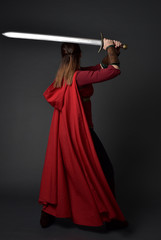 Canvas Print - full length portrait of brunette girl wearing red medieval costume and cloak. standing pose  with back to the camera, holding a sword on grey studio background.