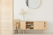 Round mirror on white wall above wooden cabinet in simple anteroom interior with armchair. Real photo
