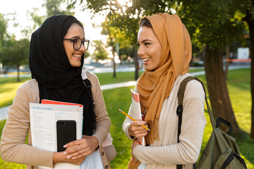 Poster - Young arabian women students holding books in park outdoors talking with each other.