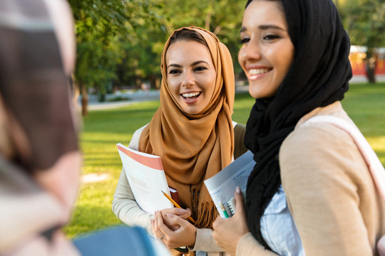 Cheerful young arabian women students holding books in park outdoors talking with each other.