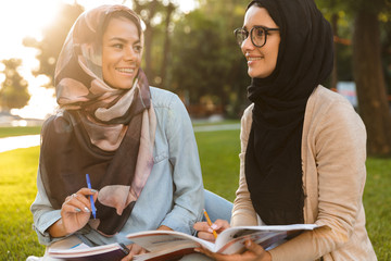 Canvas Print - Happy young arabian women students writing in copybooks in park outdoors.