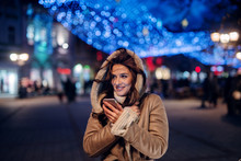 Beautiful Smiling Girl Standing Street In Winter Coat. Holding Telephone And Smiling. Christmas Decoration On Streets.Beautiful Winter Evening.