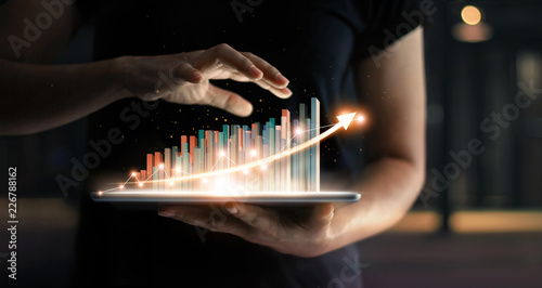 Fotomural Businessman holding tablet and showing a growing virtual hologram of statistics, graph and chart with arrow up on dark background