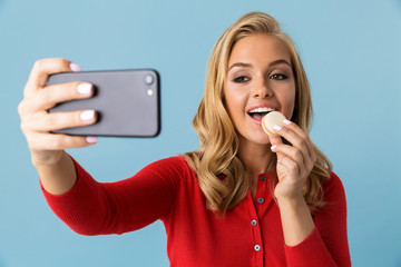 Poster - Portrait of happy blond woman 20s wearing red shirt eating biscuit and taking selfie photo on mobile phone, isolated over blue background in studio