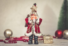 Santa Claus Toy Brings Christmas Tree At Blue Snowy Night Bokeh Background And Blurred Lights Foreground. Red Lantern Torch To Light The Way. Big Copyspace Concept New Year`s Market Banner, Poster.