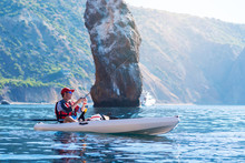 A Man Fishing On A Kayak Boat In The Sea Near The Rocks At The Shore Of Island Mountain. Young Fisherman Kayaking.