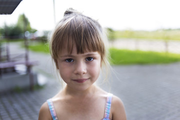 Close-up portrait of pretty young little blond pale unhappy moody friendless child girl looking sadly in camera on blurred sunny outdoors background. Children problems concept.