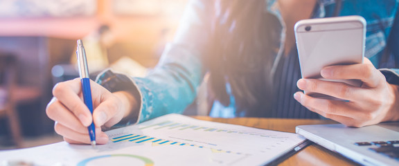 Wall Mural - Woman hand using a smartphone and writing on charts and graphs that show results with a pen in the office.Web banner.