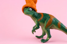 Funny Green Dinosaur Toy In Little Knitted Orange Hat  Near On Pastel Pink Background