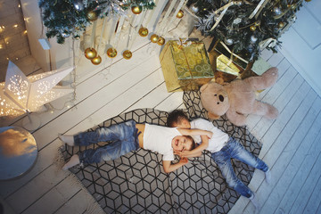 Wall Mural - Two boys in white T-shirts and blue jeans are lying on the checkered carpet near the Christmas fir. View from above.