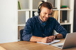 © LIGHTFIELD STUDIOS - portrait of smiling man in headphones making notes while taking part in webinar at tabletop with laptop in office