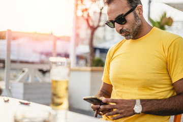 One young man playing with his smartphone while drinking a beer outdoor. Yellow t shirt dressing.