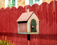 Mailbox In The Form Of A Small House On The Background Of A Red Fence