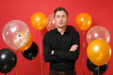 Shocked young man in black classic shirt holding hands folded on bright red background air balloons. St. Valentine's, International Women's Day, Happy New Year, birthday mockup holiday party concept.