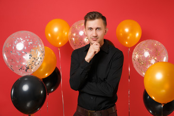 Smiling young man in black classic shirt keeping hand on chin celebrating on red background air balloons. Valentine's, International Women's Day, Happy New Year, birthday mockup holiday party concept.