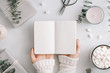 Girl's hands hold an blank notebook among gift boxes and winter festive decor. The concept of diary, holiday greetings or planning. Flat lay, top view, mockup.