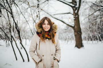 Wall Mural - Theme is a weekend holiday in winter. Beautiful young Caucasian woman standing in snow park in jacket with hood and fur in jeans and smiling