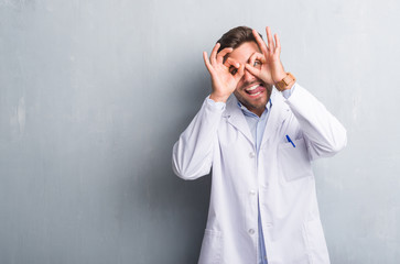 Handsome young professional man over grey grunge wall wearing white coat doing ok gesture like binoculars sticking tongue out, eyes looking through fingers. Crazy expression.