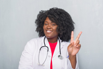 Poster - Young african american doctor woman over grey grunge wall smiling with happy face winking at the camera doing victory sign. Number two.