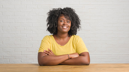 Sticker - Young african american woman sitting on the table at home happy face smiling with crossed arms looking at the camera. Positive person.