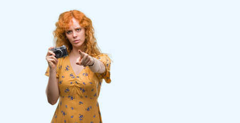 Sticker - Young redhead woman taking pictures holding vintage camera pointing with finger to the camera and to you, hand sign, positive and confident gesture from the front