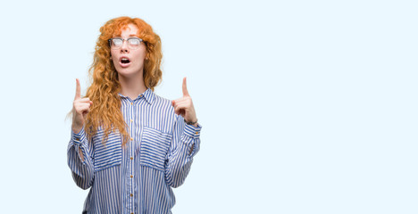 Poster - Young redhead bussines woman amazed and surprised looking up and pointing with fingers and raised arms.