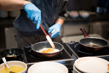 Hotel chef hands with gloves cooking fried eggs on hot pan for breakfast in restaurant at hotel.