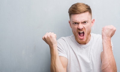 Wall Mural - Young redhead man over grey grunge wall annoyed and frustrated shouting with anger, crazy and yelling with raised hand, anger concept