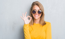 Beautiful Young Woman Standing Over Grunge Grey Wall Wearing Retro Sunglasses Doing Ok Sign With Fingers, Excellent Symbol