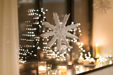 christmas and holidays concept - paper snowflake decoration hanging over garland lights and candles at night window