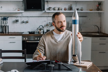 Handsome Engineer Looking At Rocket Model And Making Notes At Home