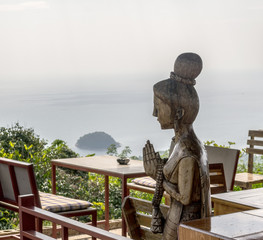 Сarved wooden sculpture of a woman welcoming in a cafe. Thai greeting wai. Beautiful seaview from the table in the restaurant.