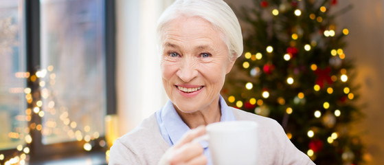 Sticker - age, holidays and people concept - happy smiling senior woman with cup of coffee at home over christmas tree lights background