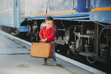 Adorable Little Kid Boy Dressed In Red Sweater On A Railway Station Near Train With Retro Old  Brown Suitcase. Ready For Vacation. Young Traveler On The Platform.
