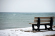 Winter/fall. The photo of the bench on the ocean cost with a bit of snow on the ground. The blue water of the ocean is on the background.
