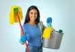 Young attractive woman holding cleaning tools and products in bucket isolated on blue background