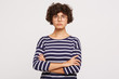 Closeup of calm serious emotionless young curly brownhair woman wears white and blue stripped sweatshirt and transparent black-framed round glasses keeps arms crossed isolated over white background