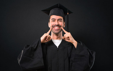 Man on his graduation day University smiling with a happy and pleasant expression while pointing mouth and face with fingers on black background