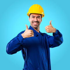 Young workman with helmet giving a thumbs up gesture and smiling because has had success on blue background