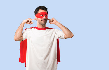 Wall Mural - Superhero man with mask and red cape covering both ears with hands on isolated blue background