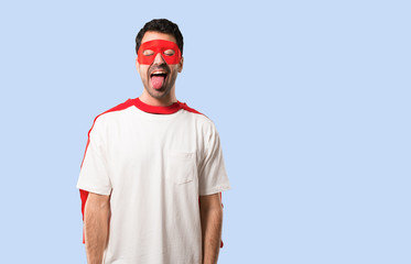 Wall Mural - Superhero man with mask and red cape showing tongue at the camera having funny look on isolated blue background