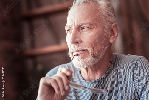 Positive thinking. Close up of elderly man keeping his glasses rim an his chin and looking away while being pleased