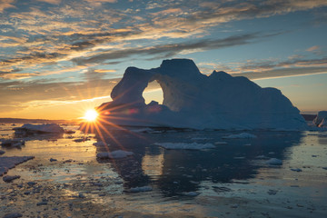 Wall Mural - sunburst behind a massive iceberg with a hole in it during the midnight sun season. disko bay, green