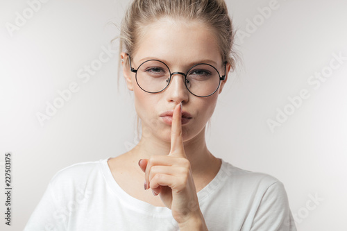 Fotografie, Obraz Cute mysterious girl wearing eyeglasses in casual outfit showing silence sign ho