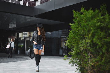 Wall Mural - Beautiful, sexy and fashionable brunette model girl with bright makeup, in the jeans shorts, in a leather jacket and in a hat, walking outdoors with a stylish black backpack in her hand